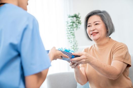 A young caregiver hand over to her senior patient a blue gift box with blue ribbons at a contented living room.