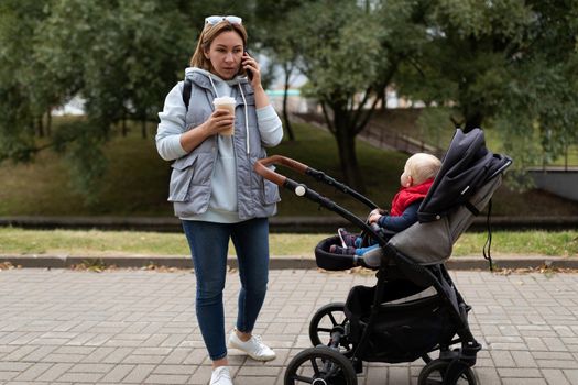 young woman while walking with her baby and pram talking on a mobile phone and drinking coffee in a city park.