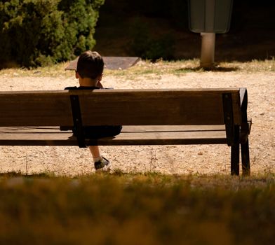 Kid sad alone in the dark at night under a streetlight sitting in a park bench. Missing his family.