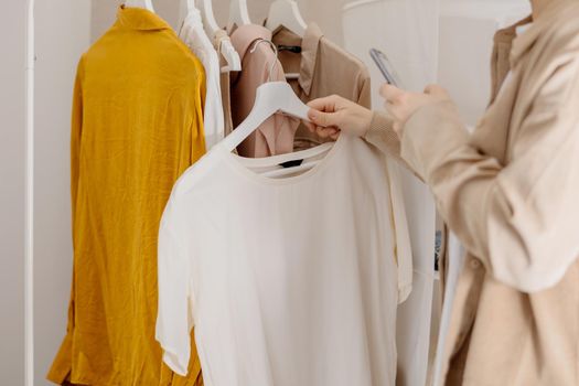 Woman holding smartphone and taking photo of her old clothes to sell them online. Selling on website, e-commerce. Reuse, second-hand concept. Conscious consumer, sustainable lifestyle. Close-up view