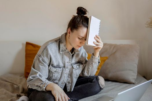 Young beautiful woman with casual clothes sitting on the bed at home with laptop computer and studying. Girl is sad and tired. Negative emotions, stress, mental problems, deadline. Distance education