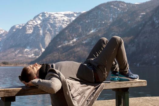 Young man lying outdoors on bench and enjoying mountains, snow, good weather, blue sky, sun. Beautiful landscape. Time with yourself, dreaming, relaxation, mental health. Tourism, holiday, travel