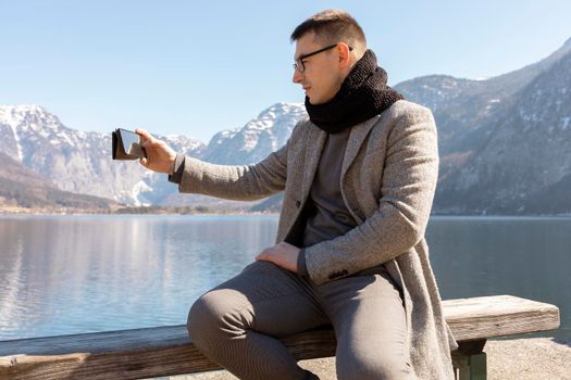 Young adult man sitting outdoors on bench and making selfie with his smartphone, enjoying mountains, lake, good weather, blue sky, sun. Beautiful, amazing landscape. Holiday, vacation, travel time