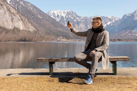 Young adult man sitting outdoors on bench and making selfie with his smartphone, enjoying mountains, lake, good weather, blue sky, sun. Beautiful, amazing landscape. Holiday, vacation, travel time