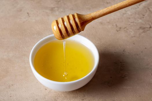 A bowl of honey and a wooden spoon for honey on a light surface. Liquid honey drips into the bowl. Selective focus.