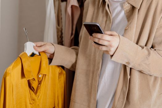 Woman holding smartphone and taking photo of her old clothes to sell them online. Selling on website, e-commerce. Reuse, second-hand concept. Conscious consumer, sustainable lifestyle. Close-up view