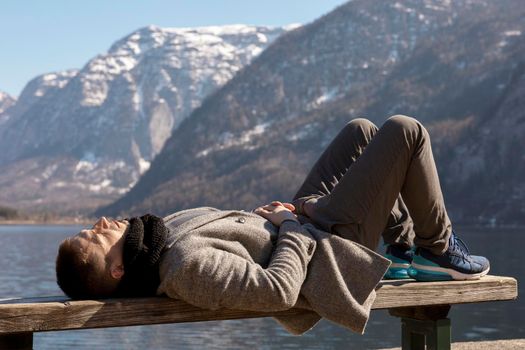 Young man lying outdoors on bench and enjoying mountains, snow, good weather, blue sky, sun. Beautiful landscape. Time with yourself, dreaming, relaxation, mental health. Tourism, holiday, travel