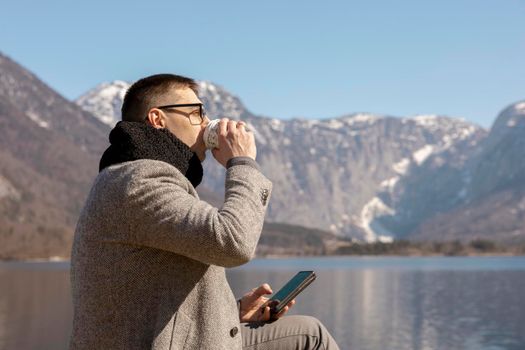 Young adult man sitting outdoors, drinking coffee and enjoying mountains, lake, good weather, blue sky and sun. Beautiful landscape. Time with yourself, dreaming, relaxation, mental health. Holiday