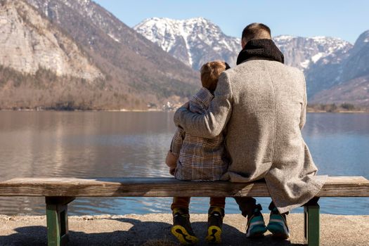 Father and son sitting together outdoors on the bench and enjoying mountains, snow, good weather, blue sky. Little boy and his father spending time together. Family time. Beautiful winter landscape