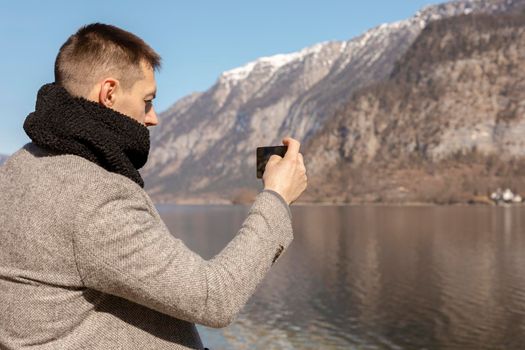 Young adult man taking picture with his smartphone, enjoying mountains, lake, good weather, blue sky, sun. Beautiful, amazing landscape. Tourism, holiday, vacation, travel time. Tourist making photo