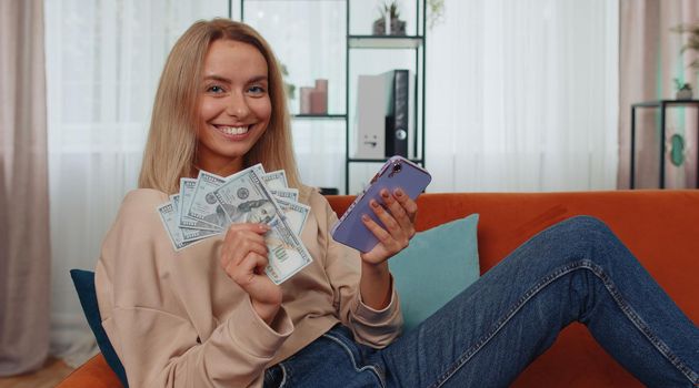 Planning family budget. Smiling caucasian girl counting money cash, use smartphone calculate domestic bills at home. Joyful young woman satisfied of income and saves money for planned vacation, gifts