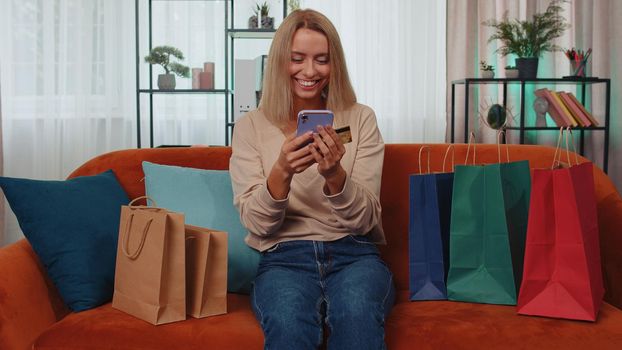 Happy shopaholic consumer girl sitting with shopping bags at modern home apartment making online order, payment with credit plastic bank card. Young woman on couch at home. Discount sale on internet