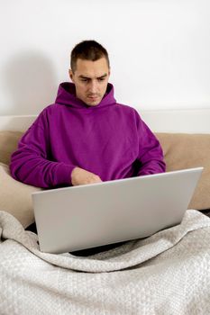 Young caucasian man with violet hoodie sitting on bed and holding laptop computer. Man using notebook to surf in internet, read news, watch movie, study or work online. Relaxing at home