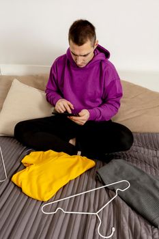 Man with violet hoodie using digital tablet, takes a photo of his old clothes to sell them online. Selling on website, e-commerce. Reuse second-hand concept. Conscious consumer, sustainable lifestyle