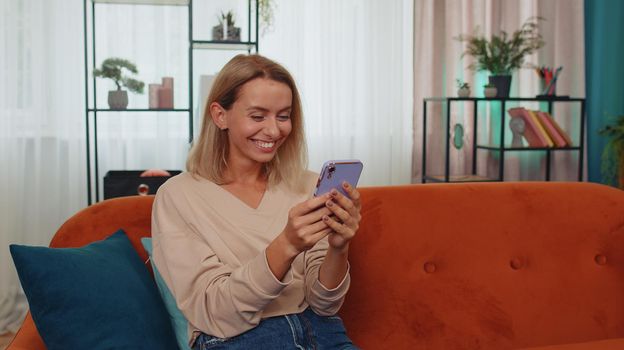 Portrait of lovely winner girl use mobile smartphone typing browsing say Wow yes found out great big win good news celebrate, lottery jackpot. Young woman at home in living room sitting on orange sofa
