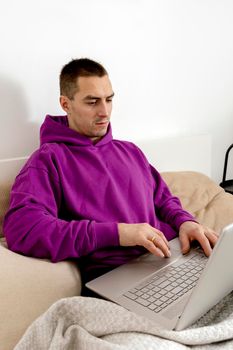 Young caucasian man with violet hoodie sitting on bed and holding laptop computer. Man using notebook to surf in internet, read news, watch movie, study or work online, call friends. Relaxing at home