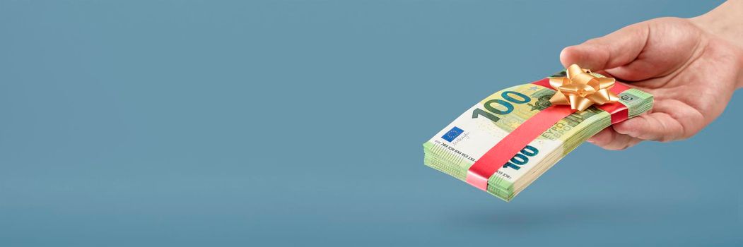 Gift of money. A man holds a stack of banknotes in his hands as a symbol or concept of a gift. 100 euro banknotes tied with a red ribbon with a bow on a blue background