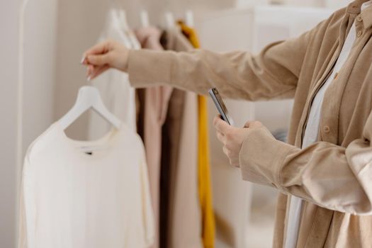 Woman holding smartphone and taking photo of her old clothes to sell them online. Selling on website, e-commerce. Reuse, second-hand concept. Conscious consumer, sustainable lifestyle. Close-up view