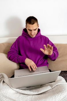Young caucasian man with violet hoodie sitting on bed and holding laptop computer. Man using notebook to study online, call friends. Distance learning, videocall, say hello. Relaxing at home