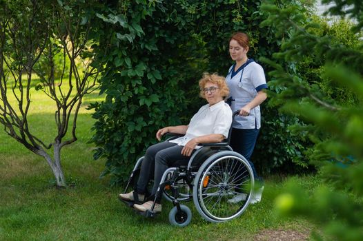 Caucasian female doctor walks with an elderly patient in a wheelchair in the park. Nurse accompanies an old woman on a walk outdoors