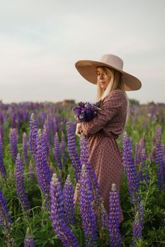 A beautiful woman in a straw hat walks in a field with purple flowers. A walk in nature in the lupin field.