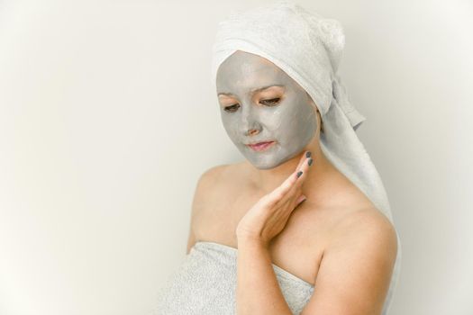 Young woman with cosmetic mask made of gray clay, wrapped in towel, touch face with hand, her gaze is directed downward. Facial skin care with cosmetic treatment.