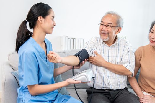 An elderly man having a blood pressure check by his personal caregiver with his wife sitting next to him in their home.