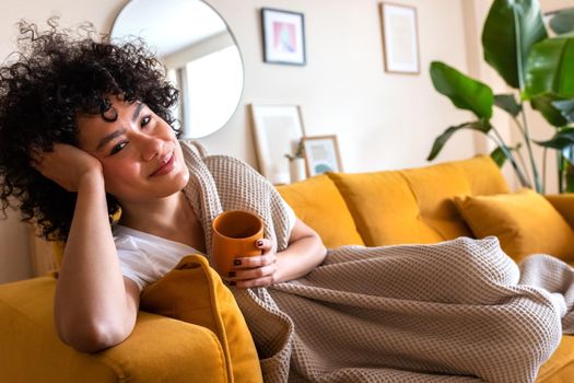 Happy, smiling African American woman relaxing at home lying on the couch holding cup of warm coffee looking at camera. Copy space. Lifestyle concept.