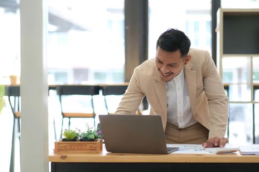 Young Asian businessman in suit working with laptop computer in office. Man in suit using laptop in well-lit workplace..