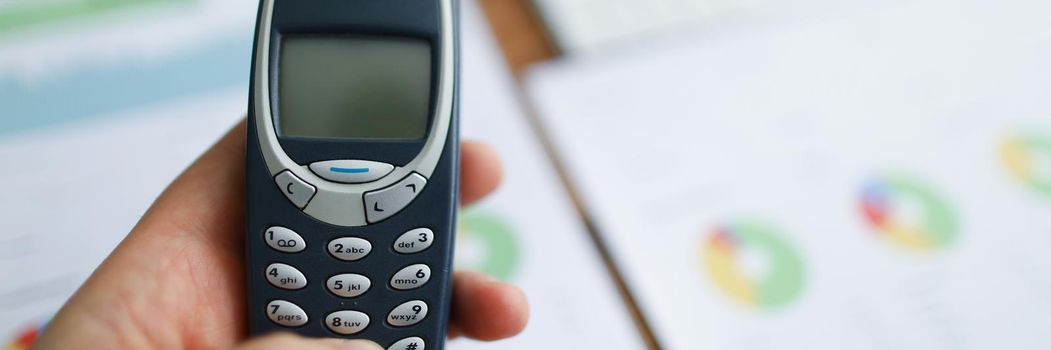 In the hands of an old model of a mobile phone against the background of reports on the table, close-up, blurry. Corporate cell phone