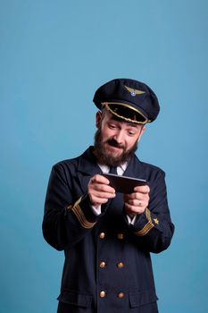 Airplane captain in professional uniform playing mobile games on smartphone, holding phone. Pilot with excited facial expression using aviation simulator on telephone, leisure entertainment