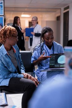 Nurse and patient analyzing brain scan on laptop, talking about tomography diagnosis and neural system on computer. Young woman and medical assistant doing checkup consultation in waiting room lobby.