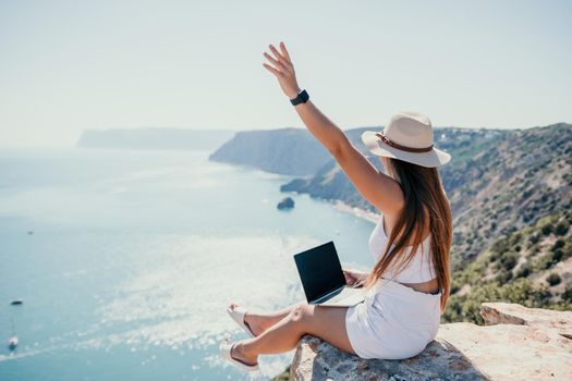 Successful business woman in yellow hat working on laptop by the sea. Pretty lady typing on computer at summer day outdoors. Freelance, travel and holidays concept.