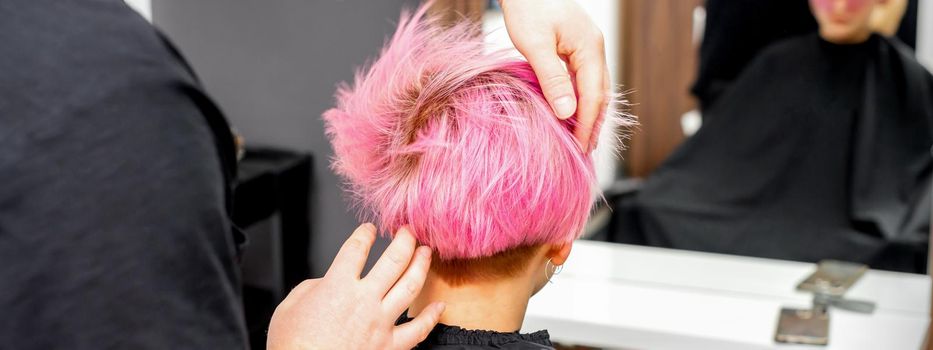 Hairdresser with hands is checking out and fixing the short pink hairstyle of the young white woman in a hair salon