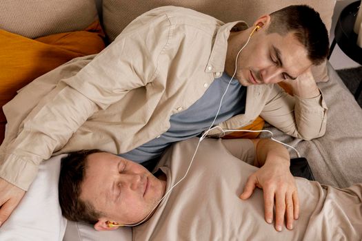 Happy gay couple with casual clothes spending time together at home and listening music with earphones. Two caucasian men relaxing. Homosexual relationships and alternative love. Cosy interior