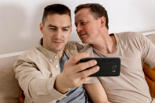 Happy gay couple with casual clothes spending time together at home and making selfie on smartphone. Homosexual relationships and alternative love. Cosy interior
