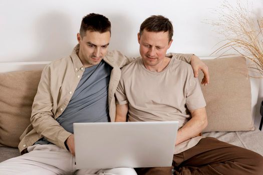 Happy gay couple with casual clothes spending time together at home and watching movie on the laptop. Two caucasian men relaxing. Homosexual relationships and alternative love. Cosy interior