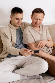 Happy gay couple with casual clothes spending time together at home and watching something on smartphone, or having video call with friends. Homosexual relationships and alternative love