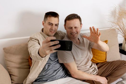 Happy gay couple with casual clothes holding smartphone and talking with friends online, having video call. Homosexual relationships and alternative love
