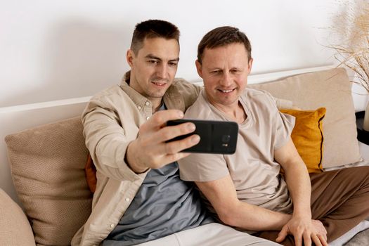 Happy gay couple with casual clothes spending time together at home and making selfie on smartphone. Homosexual relationships and alternative love. Cosy interior