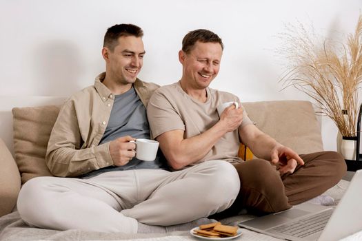 Happy gay couple with casual clothes spending time together at home, watching movie on the laptop and drinking tea. Two caucasian men relaxing. Homosexual relationships and alternative love