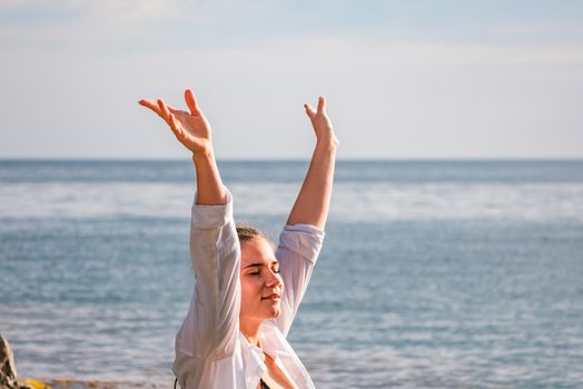 The girl stands on the shore and looks at the sea. Her hands are raised up. She wears a white shirt and her hair is in a braid