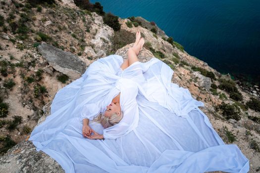 The blonde lies on a rock above the sea. With long hair on a sunny seashore in a white dress, rear view, silk fabric flutters in the wind. Against the backdrop of blue skies and mountains