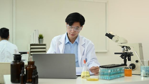 Young male Biochemist using laptop computer, collecting data and summarising information for scientific report.