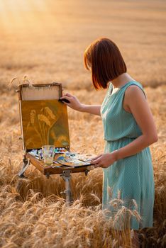 Back view of a young girl painter making beautiful still life painting of three spikelets of rye with a background of big field at sunset