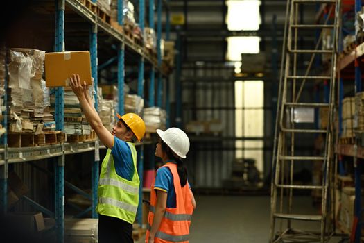 Two young colleagues wearing hardhat and reflective jacket checking stock on the shelves in a large warehouse.