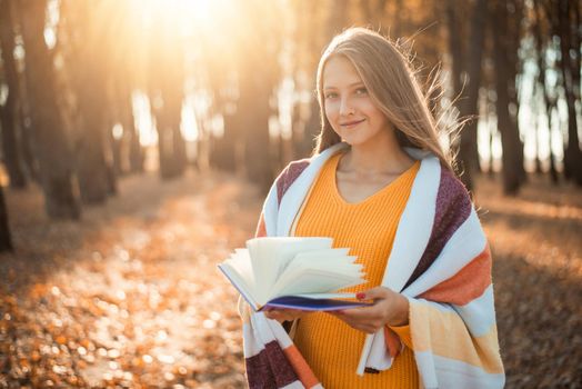 Atmospheric photo of a blond girl in warm plaid and orange sweater with a book of poems in hands in the park