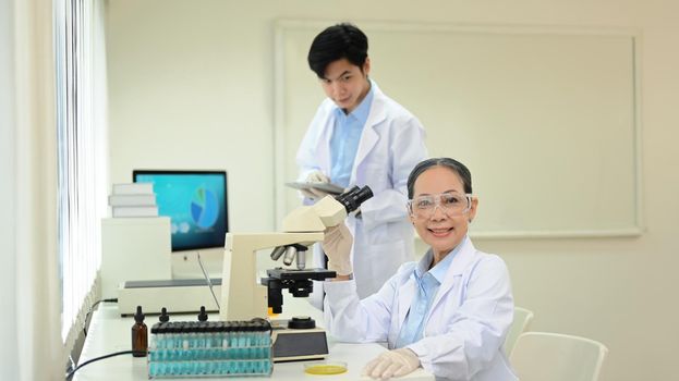Experienced senior woman scientist in white coat sitting front of microscope and smiling to camera.