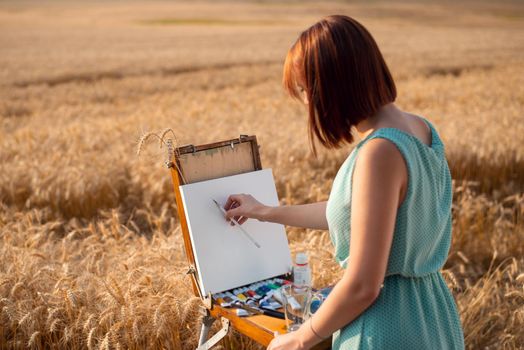 Young girl painting landscape of a field of rye at sunset