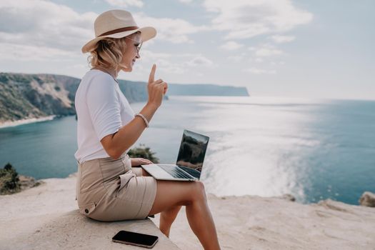 Successful business woman in yellow hat working on laptop by the sea. Pretty lady typing on computer at summer day outdoors. Freelance, travel and holidays concept.